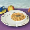 A plate with cereal and a special rim to help keep food on the plate, with a cup and an apple in the background. Produktbild-Vorschau