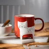 A red mug with a white cat design is placed on a marble coaster on a wooden table. A white bowl with a wooden spoon is to the left of the mug. Produktbild-Vorschau