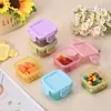 A collection of small, colorful, square plastic food storage containers filled with various snacks and candies, arranged on a light-colored surface with flowers in the background. Produktbild-Vorschau