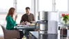A couple is having breakfast at a table with a coffee maker in the foreground. Produktbild-Vorschau