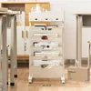 A mobile storage rack with multiple shelves and drawers is placed next to a desk and a cabinet in an office or study environment. Produktbild-Vorschau