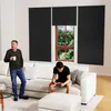 Two men in a living room, one standing with a mug and the other sitting on a sofa reading a book. Black roller blinds are installed on the window. Produktbild-Vorschau