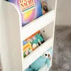 A white, multi-tiered shelf unit holding children's books and a stuffed toy. The shelf is placed on a gray carpeted floor. Podgląd zdjęcia produktu