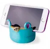A smartphone is held upright by a blue, cartoon-like phone stand. Produktbild-Vorschau