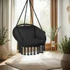 A black hanging chair with fringe detail is shown in a well-lit room with large windows, plants, and a rug. Náhled obrázku produktu