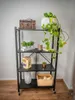 A black metal shelving unit with various decorative items on its shelves, placed against a light gray wall. Produktbild-Vorschau