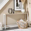 A narrow console table with a wooden top and white metal legs, placed in a hallway next to a staircase. It is decorated with a black wire lantern, a small black candle holder, a vase with dried flowers, and a woven basket filled with patterned pillows and  Anteprima dell’immagine prodotto