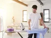 A man ironing a blue shirt in a well-lit room with a punching bag in the background. Produktbild-Vorschau