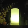 A glowing cylindrical lamp is placed on a reflective surface, with flowers and branches in the background. Produktbild-Vorschau