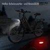 An e-bike is shown in a dark, atmospheric setting, with its headlight and taillight illuminated. The background is blurred, suggesting motion or a city environment at night. Produktbild-Vorschau