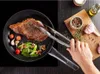 A hand is using tongs to flip a steak in a frying pan, with vegetables and seasonings nearby. Podgląd zdjęcia produktu