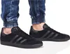 A person wearing black Adidas Gazelle sneakers and blue jeans. Voir les images du produit
