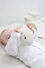 A baby is lying on a white surface and playing with a white plush whale toy. Another larger white plush whale toy is in the background. Anteprima dell’immagine prodotto