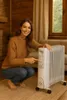 A woman is crouching next to a white oil-filled radiator in a cozy room. Podgląd zdjęcia produktu