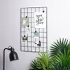 A black metal grid photo frame is mounted on a wall, displaying several small photos and a motivational quote. A black desk lamp is visible to the left, and lush green plants are in the foreground to the right. Anteprima dell’immagine prodotto