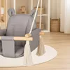 A close-up of a gray baby swing with wooden accents and tassels, placed on a white rug in a room with shelves in the background. Náhled obrázku produktu