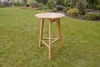 A wooden bar table is placed on a grassy lawn in a garden setting, with landscaped bushes and trees in the background. Produktbild-Vorschau
