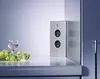 A corner electrical outlet with two sockets and two small circular indicators, placed on a white countertop next to a glass of white wine and a bunch of green grapes. Produktbild-Vorschau