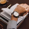 A man's wrist wearing a watch is shown over an open book, with a cup of coffee in the background. Podgląd zdjęcia produktu