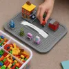 A person's hand is placing a small red figure onto a gray building block base plate. There are several small building block structures and a container of building blocks in the foreground. Produktbild-Vorschau