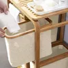 A person's hands are placing a white towel into a beige fabric laundry basket, which is part of a multi-tiered bamboo shelf unit. Podgląd zdjęcia produktu