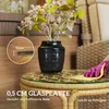 A person's hand in a pink glove is cleaning a round rattan side table with a glass top. A black vase with flowers sits on the table. Podgląd zdjęcia produktu