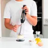 A person is using a black and silver immersion blender in a clear glass with a light-colored mixture inside. Ingredients like an egg and oil are visible on the counter. Podgląd zdjęcia produktu