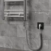 A chrome towel warmer element with a coiled power cord plugged into a black electrical outlet on a gray tiled wall. Náhled obrázku produktu