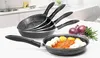 A set of frying pans is stacked on a countertop, with one pan in the foreground containing food. Red onions and rosemary are visible in the background. Náhľad obrázku produktu