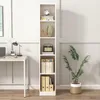 A tall, narrow white bookshelf filled with books and decorative items, placed next to a desk with a laptop and lamp, and a large potted plant. Podgląd zdjęcia produktu