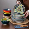 A person is holding a plate from a set of dishes, with other dishes stacked nearby on a wooden table. Náhled obrázku produktu