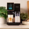 A coffee machine is shown on a countertop with a glass of coffee and a milk container. Produktbild-Vorschau