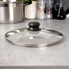 A close-up shot of a glass pot lid with a black handle, resting on a speckled gray countertop. In the background, a stainless steel pot and kitchen accessories are visible. Náhled obrázku produktu