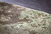 A close-up, angled view of a rug with a distressed, abstract pattern in shades of green and brown. Produktbild-Vorschau