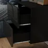 A close-up view of a black high-gloss nightstand with two drawers partially open, showing the drawer slides. The background includes a blue upholstered bed and a wooden floor. Podgląd zdjęcia produktu
