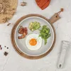 A breakfast plate with a fried egg, bacon, avocado, and greens, set on a wooden board. Produktbild-Vorschau