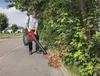 A person is using a leaf blower on a sidewalk next to a hedge. Produktbild-Vorschau