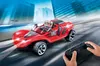 A red remote-controlled race car with a driver figure is shown speeding down a road with city buildings in the background. A child's hands are holding the remote control. Podgląd zdjęcia produktu