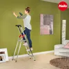 A woman is using a ladder to drill a hole in a green wall in a living room. Anteprima dell’immagine prodotto