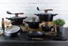 A collection of black cookware with brown handles and glass lids, arranged on a dark countertop against a white brick wall. Anteprima dell’immagine prodotto