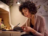 A woman is sitting at a desk, reading a book and writing in it. The desk has some plants and jars on it. There are drawings of birds on the wall behind her. Voir les images du produit