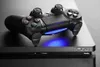 A black PlayStation 4 Slim console with a black controller on top, with a blue light emanating from the console. Produktbild-Vorschau