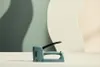 A teal stapler is placed on a white surface in front of a background with wavy green and cream shapes. Podgląd zdjęcia produktu