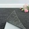 A close-up of a self-adhesive carpet tile being peeled back, revealing its backing. The background shows a floor and a white baseboard, with blurred pink flowers and a vase in the distance. Produktbild-Vorschau
