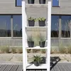 A white plant stand with several potted plants on it, set against a modern wooden building with large windows. Produktbild-Vorschau