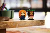 Two LEGO BrickHeadz figures, Merida and Moana, are displayed on a wooden surface. In the background, a green container holds colored pencils. Produktbild-Vorschau