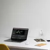 A laptop is open on a desk, displaying video editing software. A power adapter is plugged into a wall socket and connected to the laptop. Other items like a notebook and a phone are also on the desk. Produktbild-Vorschau