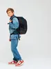 A young boy wearing a denim jacket and jeans, with a backpack on his back, stands against a white background. Produktbild-Vorschau