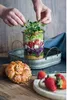 A layered salad in a glass jar being garnished by hands, with bread and strawberries in the foreground. Náhled obrázku produktu