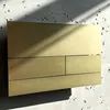 A gold flush plate for a toilet, mounted on a textured wall with sunlight casting shadows. Produktbild-Vorschau